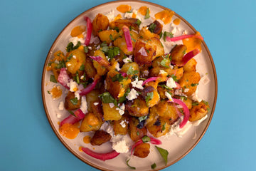 A white plate with Burnt Pineapple Hot Sauce covered roast potatoes, that is on a bed of tzatziki, and covered with pickled onion and crumbled feta.