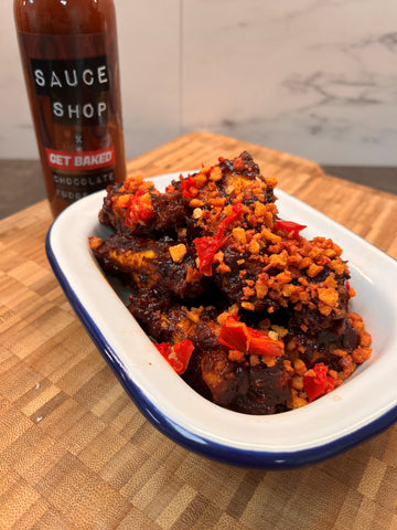 Crispy chicken wings with bacon crumbles and chilli sprinkled over the top, sat in a white bowl with Sauce Shop GET BAKED Chocolate Fudge BBQ Sauce in the background
