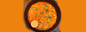 A golden coconut prawn curry in a black skillet, with a lemon on the side and an orange background that matches the Golden Coconut Prawn curry