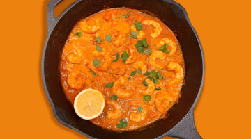 A golden coconut prawn curry in a black skillet, with a lemon on the side and an orange background that matches the Golden Coconut Prawn curry