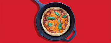 A pizza in a skillet with Sauce Shop Buffalo Hot Sauce on top and a red background