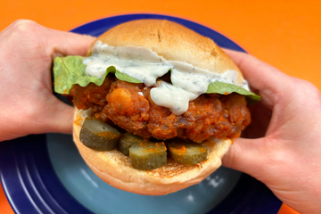 A person holding a buffalo burger up to the camera, the burger has pickles, ranch mayo, lettuce, buffalo chicken, and a brioche bun. 