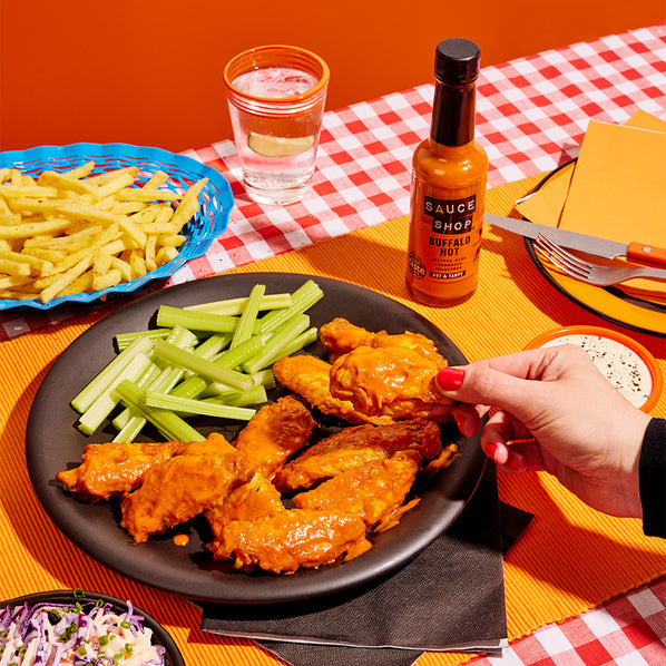 Sauce Shop Buffalo Hot Sauce on a dining table with a plate of Sauce Shop Buffalo Hot Sauce wings, fries and celery sticks. A hand is picking up a buffalo wing.