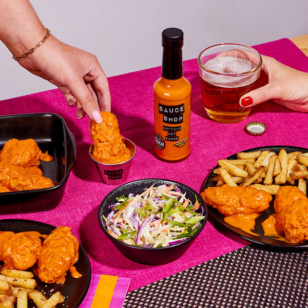 A bottle of Sauce Shop Buffalo Hot Sauce surrounded by plates of buffalo chicken wings, chips, coleslaw and beer. A hand dips a buffalo wing into a pot of Sauce Shop Buffalo Hot Sauce.
