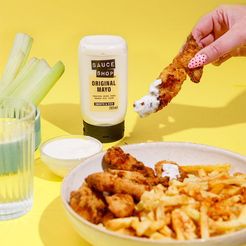  Sauce Shop Original Mayo Pack stood next to a bowl of chicken dippers and fries. A hand is picking up a chicken dipper covered in Sauce Shop Original Mayo. 